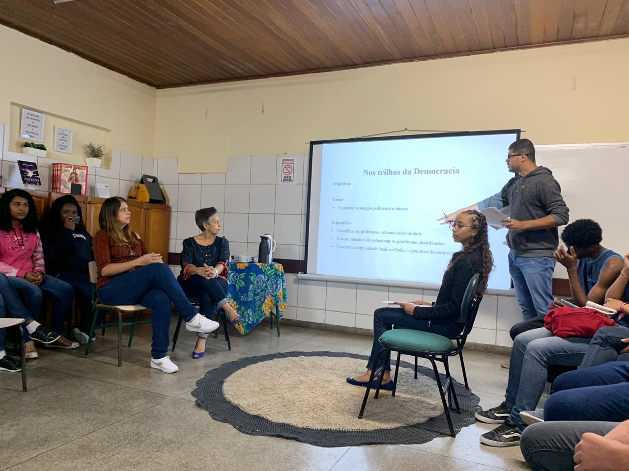 Interior de uma sala de aula com um homem em pé apontando para o quadro e possivelmente fazendo uma apresentação para outras pessoas que estão sentadas à sua frente