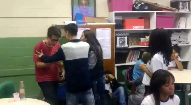 Imagem de uma sala de aula, onde parece que tem um menino sofrendo um bullyng num canto enquanto outro menino o segura com as duas mãos