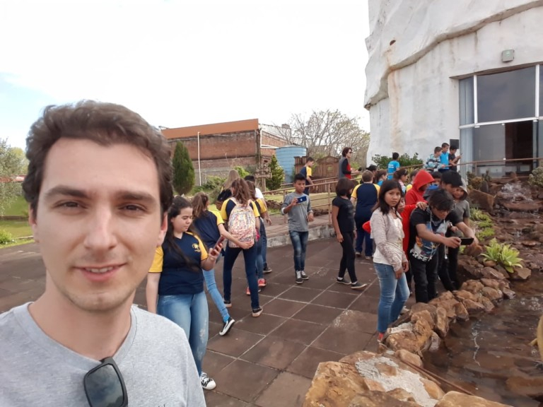 Imagem de um homem branco, franzino, com óculos de sol preso na gola da camisa. Ele parece ter feito uma selfie e ao fundo, há várias pessoas em uma área ao ar livre
