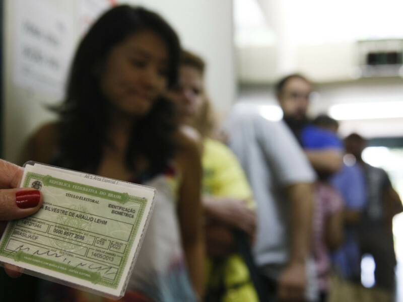 Imagem desfocada de uma fila de pessoas em algum lugar, que parece ser para votação, pois na frente há um título de eleitor nítido