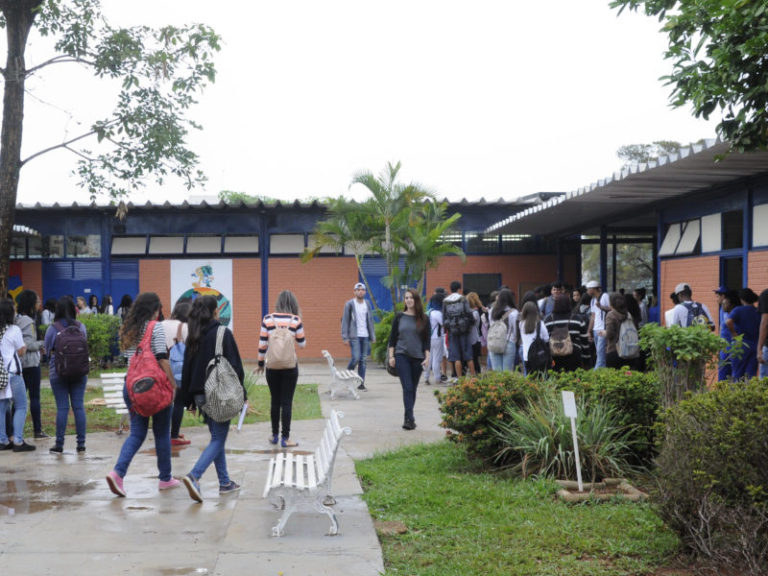 Imagem de vários estudantes chegando à porta de entrada de um colégio. É uma imagem ampla, feita ao ar livre, aprecem árvores, o céu e um pequenino jardim também