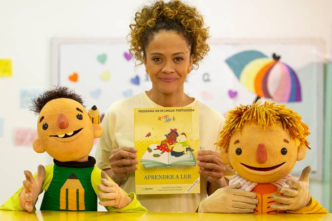 Mulher negra segurando um livro amarelo está no centro da imagem posando para a foto entre dois bonecos infantis