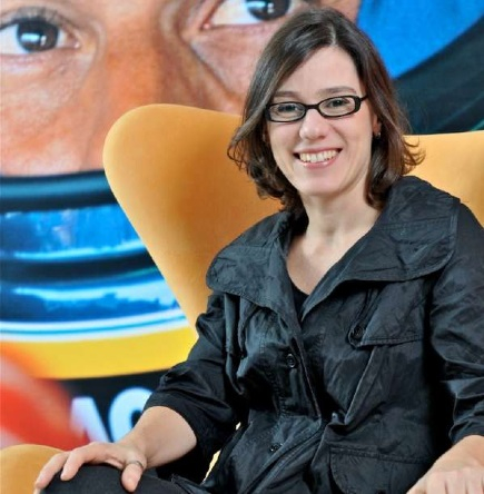 Imagem de uma mulher jovem e branca. Ela está sorridente, usando óculos de grau de armação fina e preta, um casaco de couro preto e tem cabelos curtos e castanhos, que chegam à altura do pescoço