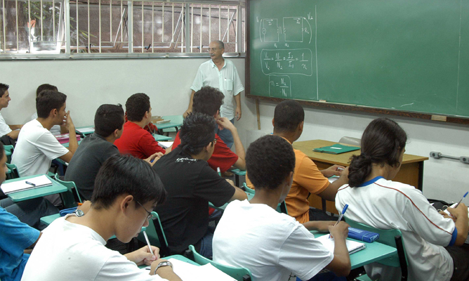 Imagem de uma sala de aula repleta de alunos sentados e escrevendo em seus cadernos ou observando o professor explicando algo na frente