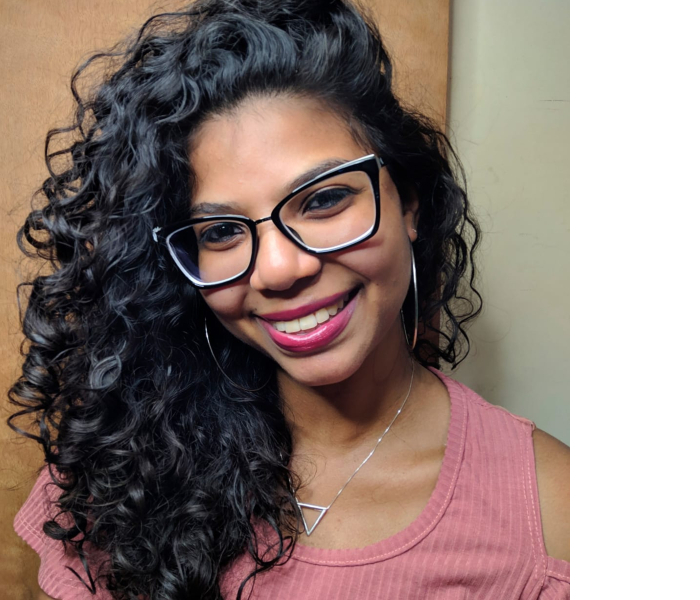 Foto de Carol, uma linda mulher negra de cabelos bem escuros e encaracolados. Ela está de blusa rosa, cordão prateado, brincos, batom rosa e óculos de grau com armação preta
