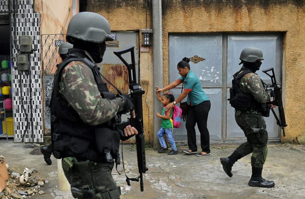 Imagem de dois militares uniformizados com o típico traje camuflado, colete à prova de balas e capacete, além de estarem segurando fuzis. Eles estão andando na rua e atrás há uma moça negra de mãos dadas com uma criança negra