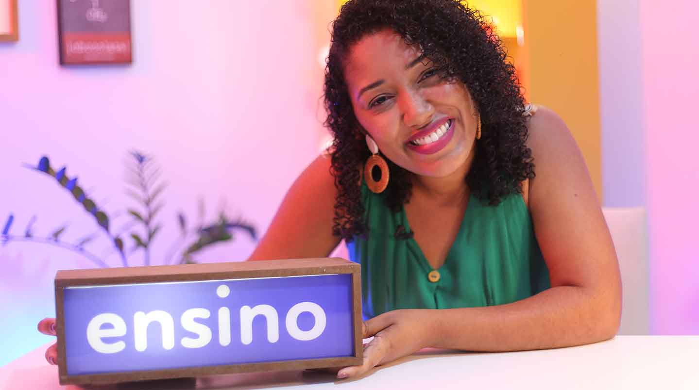 Mulher negra sorridente, de brincos de argola, camiseta verde e bem sorridente está olhando para quem bateu a foto e segurando um painel com a palavra "ensino" sendo mostrada