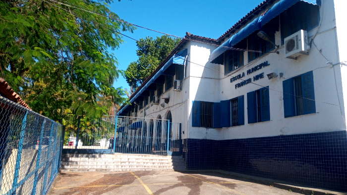 Foto da fachada de uma escola, com muros brancos e janelas azuis. 