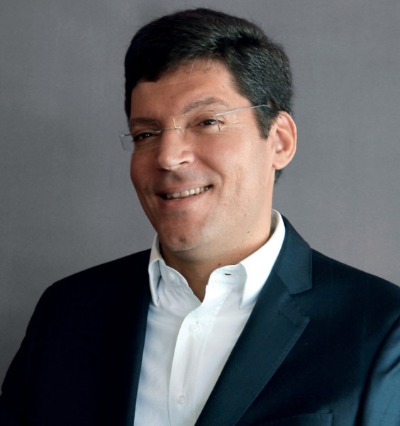 Um homem branco de cabelos pretos, curtos e bem penteados para a direita está sorrindo e posando para quem bateu a foto. Ele usa óculos e está de paletó escuro e camisa social branca por baixo do paletó