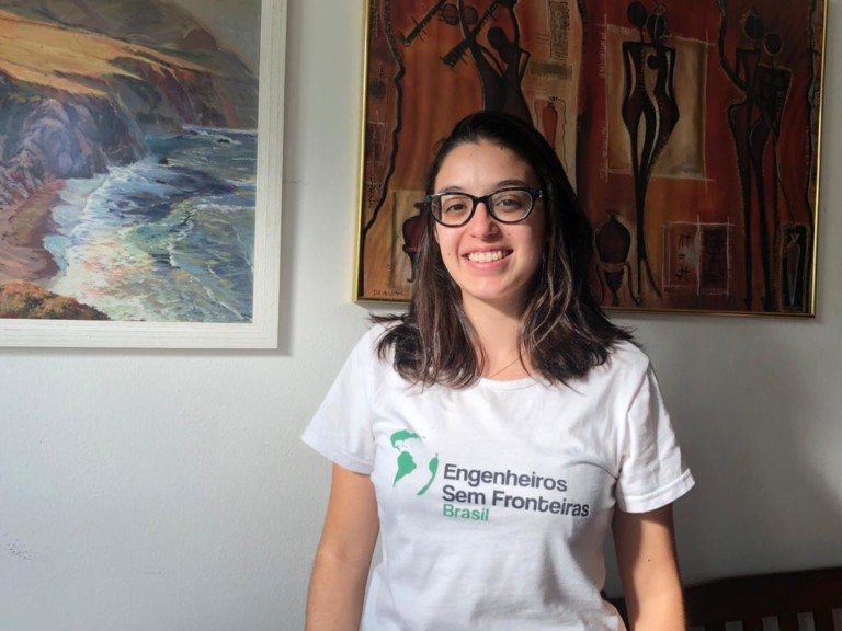 Imagem de uma jovem menina branca, sorridente, de óculos de grau e cabelos soltos. Ela esta de blusa branca com a logo do Engenheiros sem Fronteira