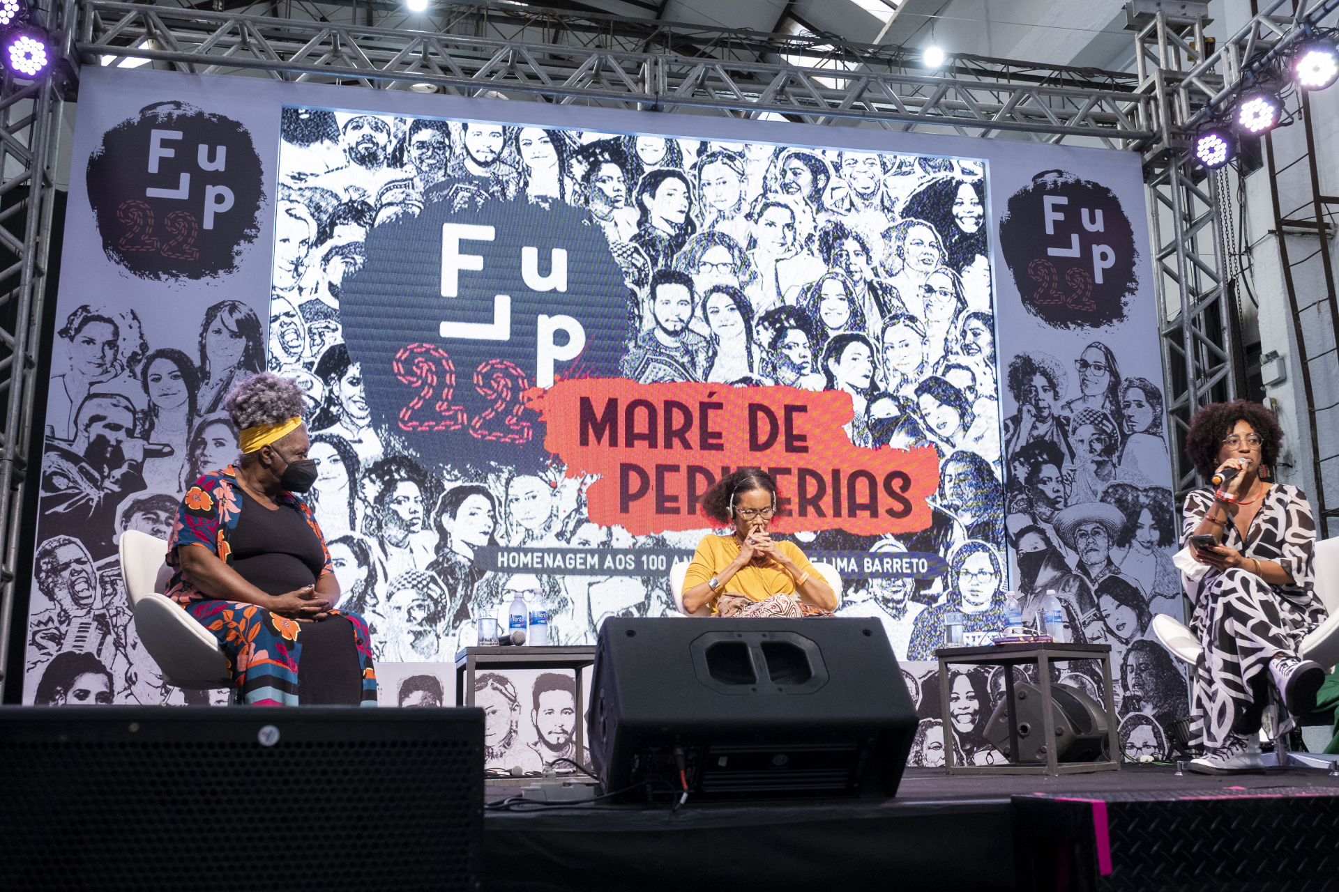 Três mulheres negras estão em um palco. Ao fundo, um grande cartaz com o nome do programa.