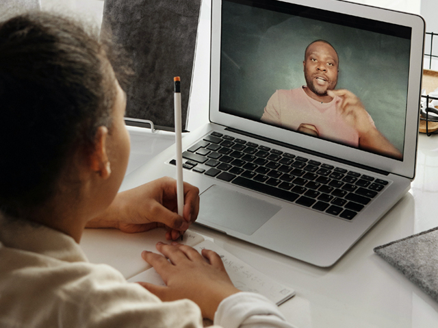 Menina negra está de costas para quem fez a foto. Ela está sentada de frente a um notebook ligado em uma videoaula. Na tela podemos ver um professor falando com ela