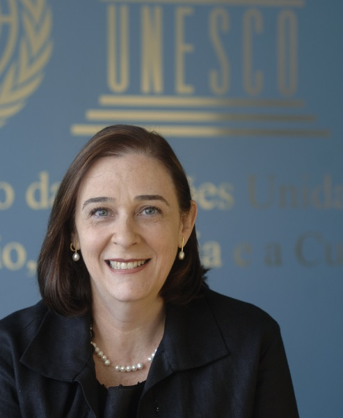 Mulher branca de meia idade está sorrindo e posando para a foto. Ela está usando um colar de pérolas, brincos elegantes e tem cabelos lisos que chegam na altura dos ombros