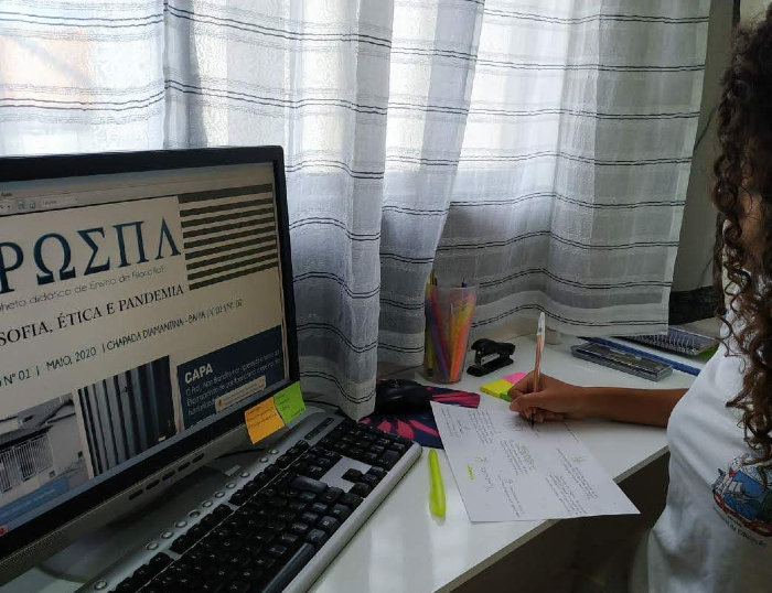 Imagem de uma menina estudado sobre filosofia em frente ao computador e escrevendo algo em uma folha