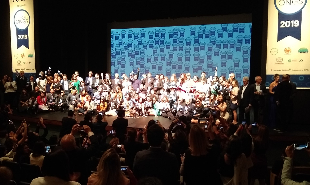 Imagem de dezenas de pessoas em um palco durante uma cerimônia de premiação. Estão felizes, alguns sentados, vários levantando os braços...podemos observar também a plateia no local