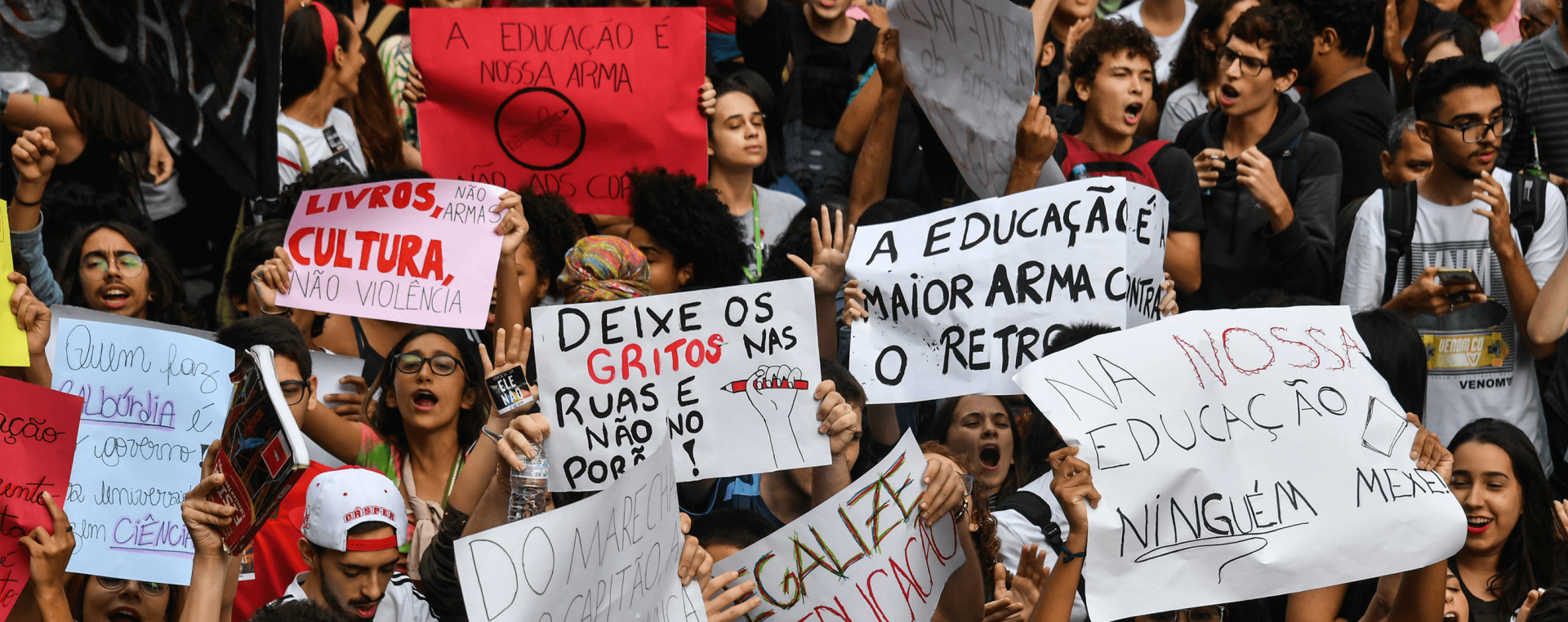 Imagem de várias pessoas segurando cartazes com dizeres de protestos ao ar livre. São protestos exigindo educação