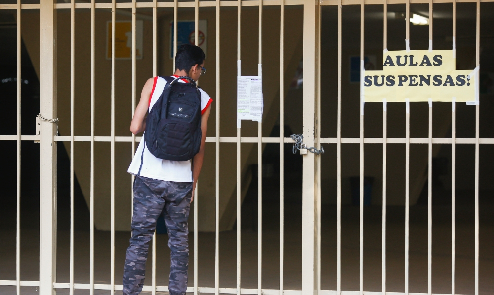Um estudante está uniformizado e de mochila às costas parado em frente à entrada do colégio, que está com as portas fechadas e um cartaz escrito "Aulas suspensas"