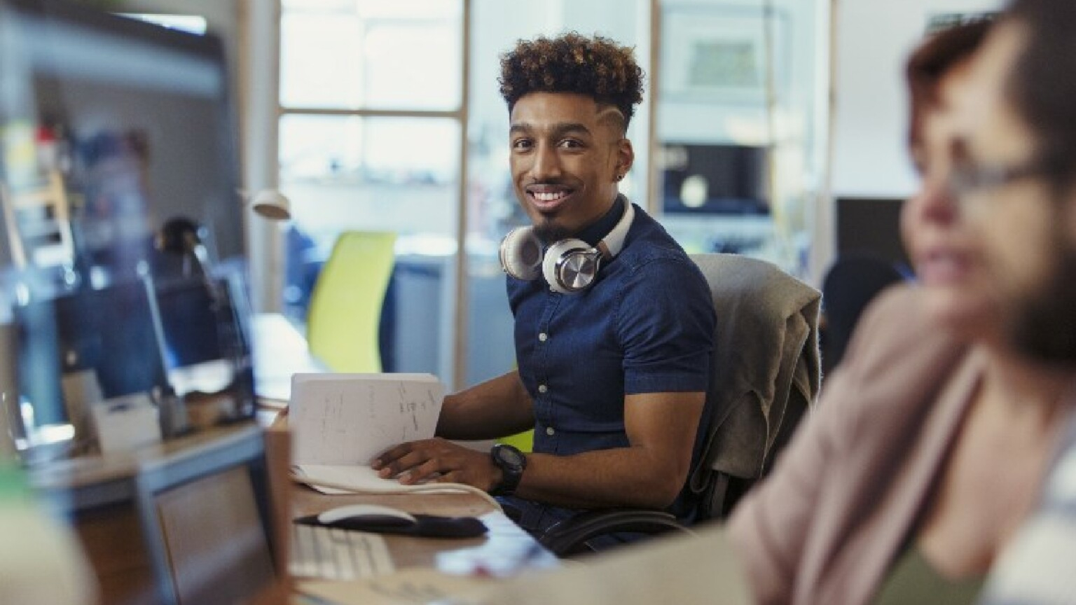 Jovem rapaz negro está de headphone pendurado no pescoço, camisa social azul marinho, com um livro aberto nas mãos e olhando sorridente para quem bateu a foto
