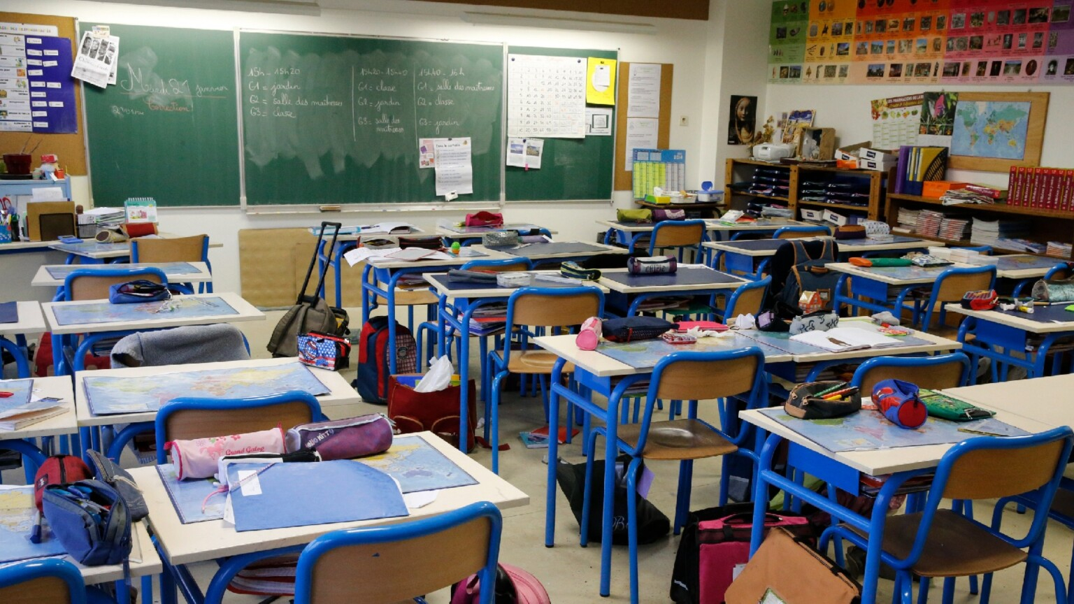 Sala de aula vazia de pessoas, com as cadeiras com materiais escolares e mochilas e o quadro negro ao fundo. Tudo indica ter sido fotografada no momento do recreio
