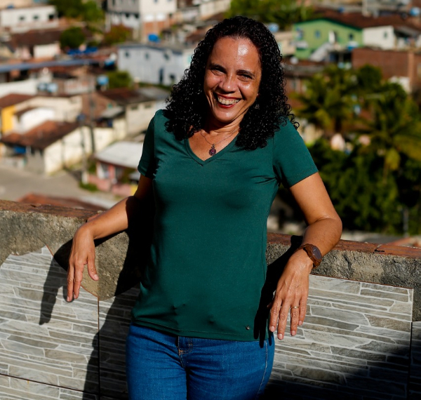 Imagem de uma mulher em pé encostada em uma marquise no alto de um prédio provavelmente, pois é possível ver, ao fundo, várias casas em um plano inferior. Ela está sorridente, tem pele morena e cabelos soltos encaracolados. Está de blusa verde escura e calça jeans
