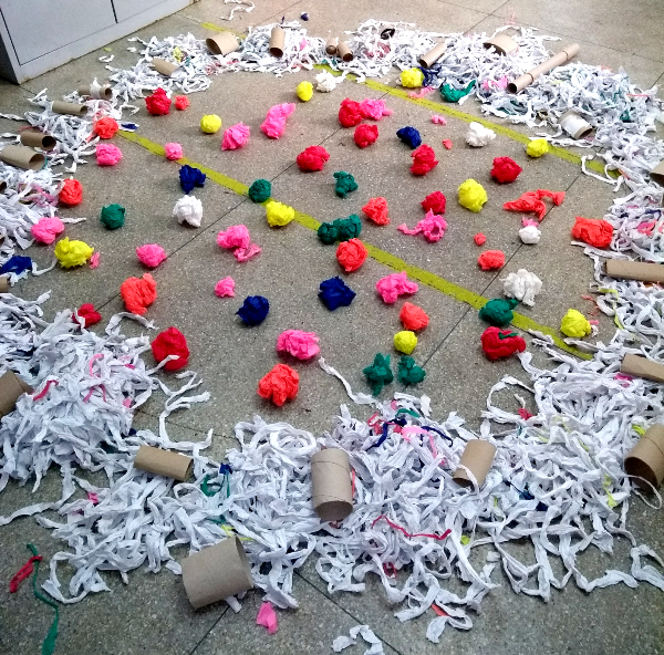 Imagem de várias papéis crepons coloridos e enrolados, formando bolinhas. Estão todos espalhados pelo chão, dentro de um círculo que foi formado por pedaços de papel branco cortados junto com rolos de papel higiênico