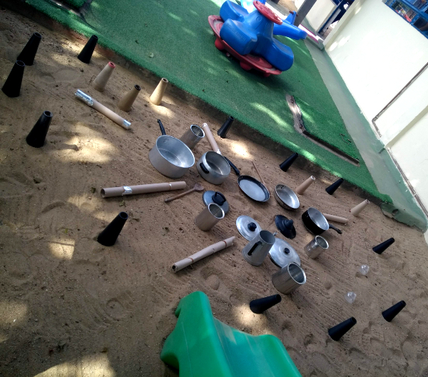 Imagem de várias panelas e suas tampas, frigideiras, rolos de linha vazios e rolos de papelão espalhados por um chão de areia