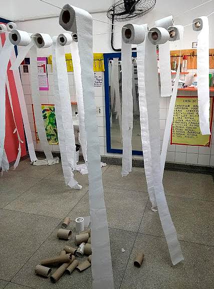 Imagem de rolos de papéis higiênicos pendurados em barbantes dentro de uma sala com vários cartazes colados na parede ao fundo.