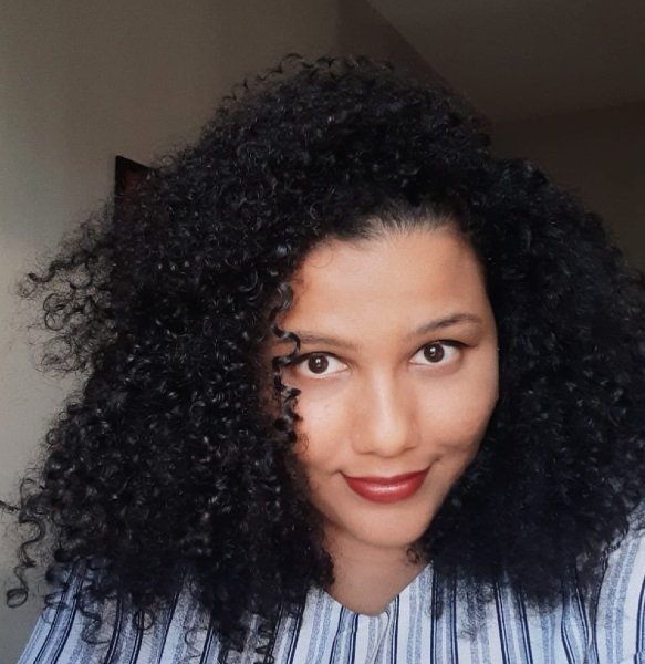 Imagem de Nataly, uma mulher com cabelos escuros, bem volumosos e encaracolados. Ela está de batom vermelho