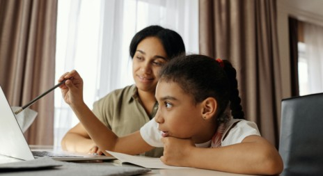 A imagem mostra uma menina negra de cabelos encaracolados, segurando um lápis com a mão direita e olhando para a tela de um computador com atenção. Ao lado dela, há uma mulher (parece ser a mãe) olhando sorridente para a filha estudando