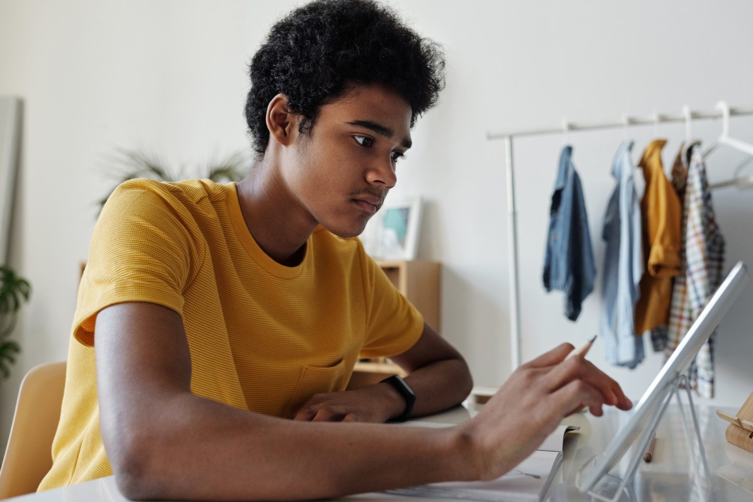 Uma pessoa negra trajando uma camisa amarela e relógio no pulso esquerdo está sentada e mexendo em um tablet branco com a mão direita