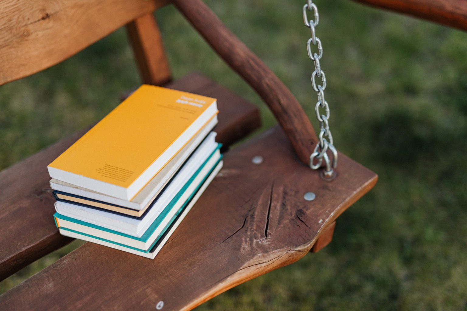 Imagem de uma pequena pilha de livros coloridos, sendo amarelo o que está no topo, está em cima de um banco de madeira suspenso por uma corrente