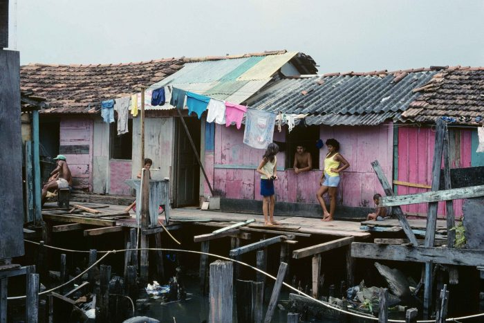 Imagem de uma casa de madeira bem humilde. É uma casa na cor de rosa e com um varal de roupas pendurado do lado de fora. Na imagem podemos perceber que a casa foi construída sobre a água e há crianças e adultos interagindo do lado de fora, trajando roupas humildes