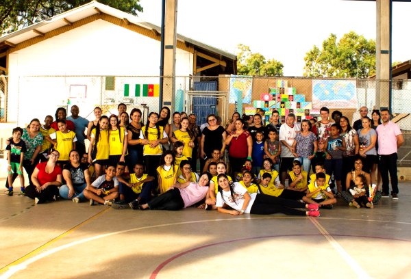 Imagem de várias crianças uniformizadas ao lado de professores. Todos estão posando para a foto em uma quadra poliesportiva que parece estar ao ar livre. Atrás das pessoas há um mural com bandeiras de países africanos, máscaras africanas e um mapa do continente