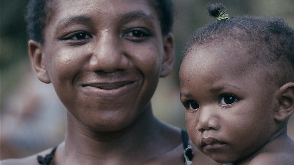 Uma mulher negra sorrindo sem mostras os dentes está com um bebê no colo. A imagem está mostrando apenas o rosto de ambas