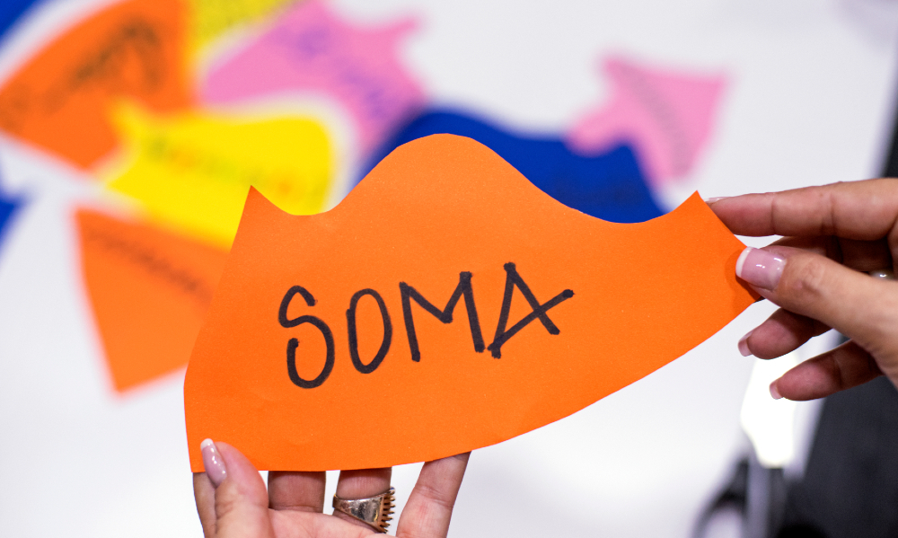Imagem de duas mãos de uma mulher adulta branca segurando um papel de cor laranja com a palavra "soma" escrito no centro