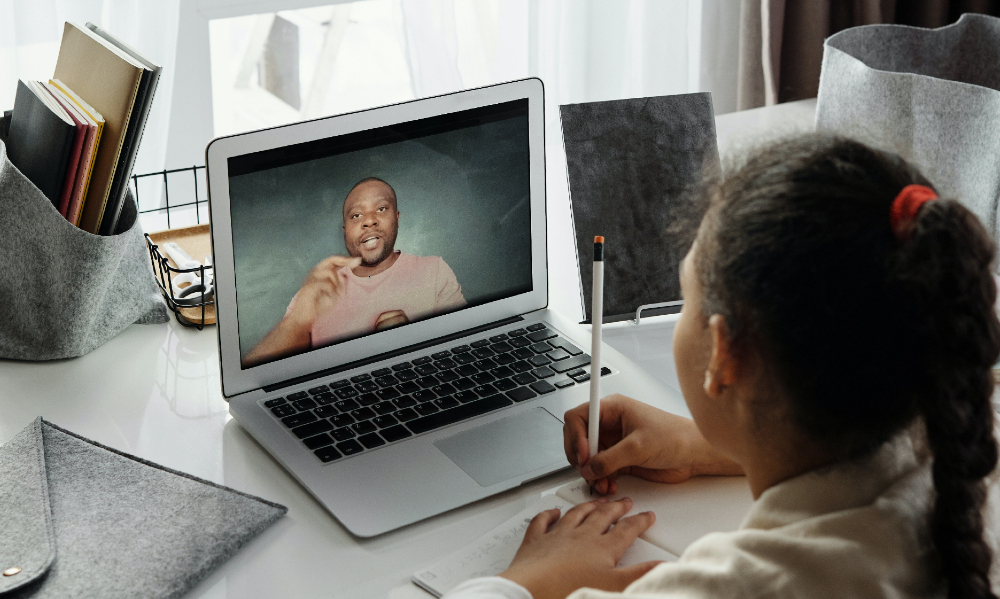 Jovem menina negra está assistindo uma videoaula pelo notebook. Na tela do computador podemos observar um professor falando algo