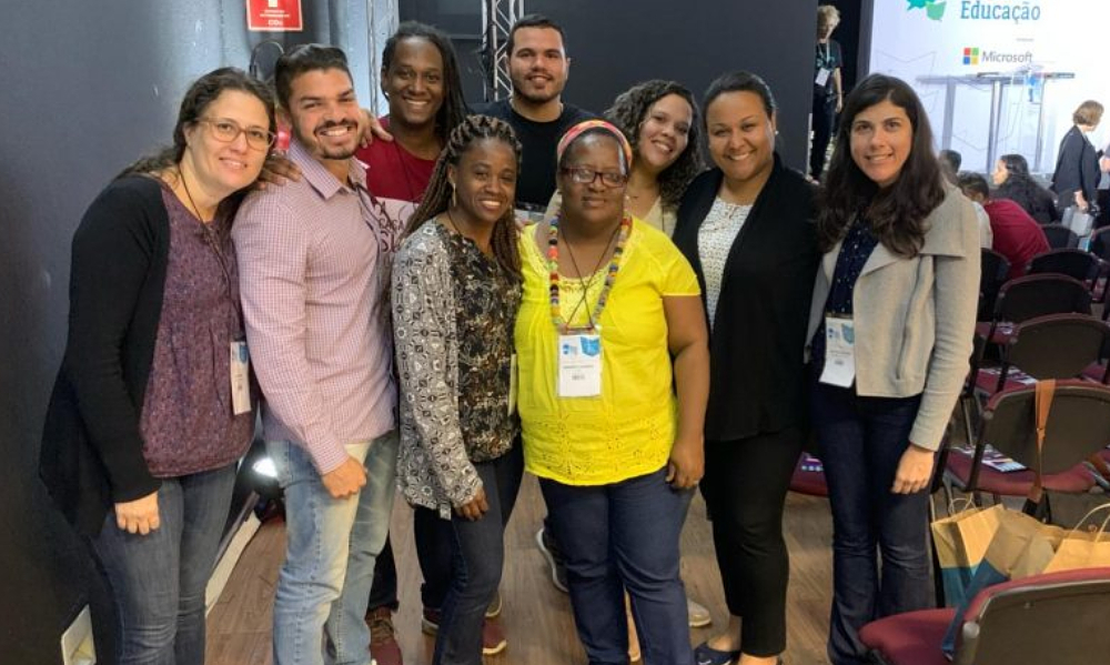 Nove pessoas em pé posando para a foto lado a lado. Estão sorridentes e são seis mulheres e três homens