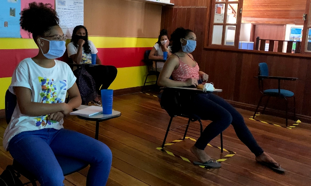 Quatro mulheres estão sentadas em cadeiras de uma sala de aula, separadas por uma distância razoável e usando máscara de prevenção à covid
