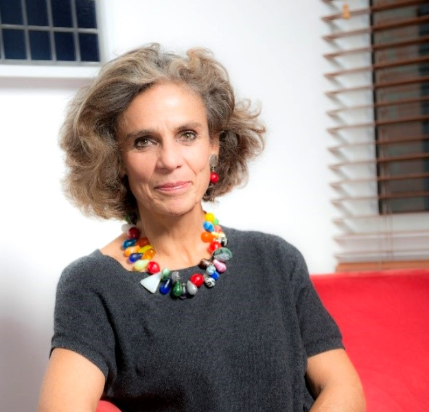 Uma mulher branca, bem maquiada, que parece ter por volta de 50 anos, está olhando para quem bateu a foto e sorrindo sem mostrar os dentes. Ela usa uma blusa cinza e um belo colar com dezenas de pedras coloridas