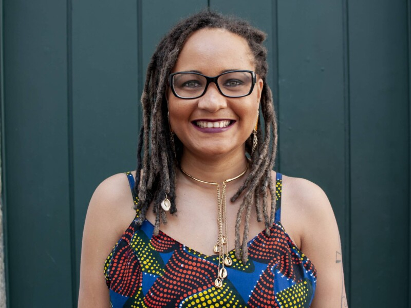 Mulher negra de cabelo rastafari, óculos de grau, colar e camiseta colorida está sorrindo para quem bateu a foto