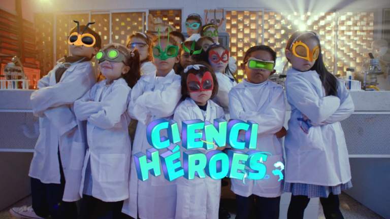 Uma dezena de crianças usando jalecos brancos e óculos coloridos posando para a foto, com a logomarca do programa Cienciheroes à frente