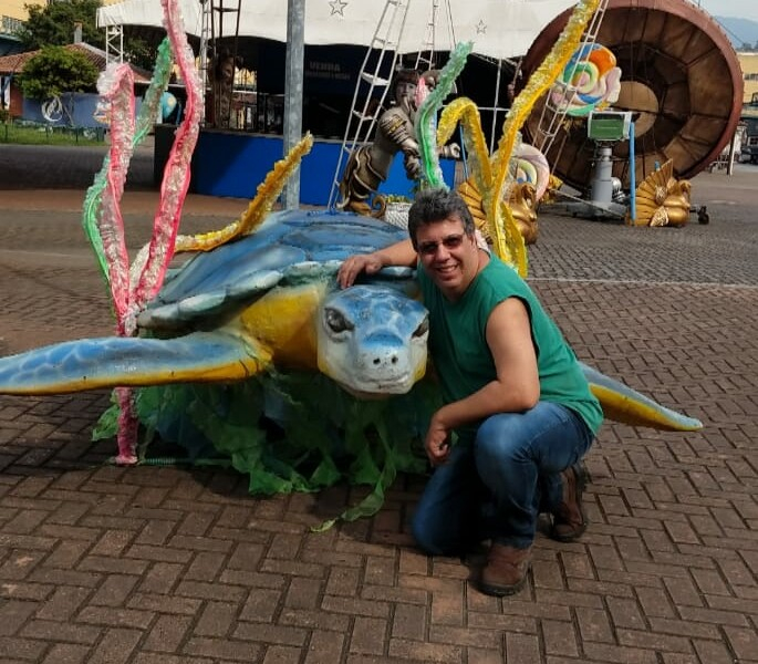 Um homem branco de óculos de sol está agachado e abraçando uma tartaruga azul e amarela, que é na verdade uma alegoria de um carro alegórico de samba