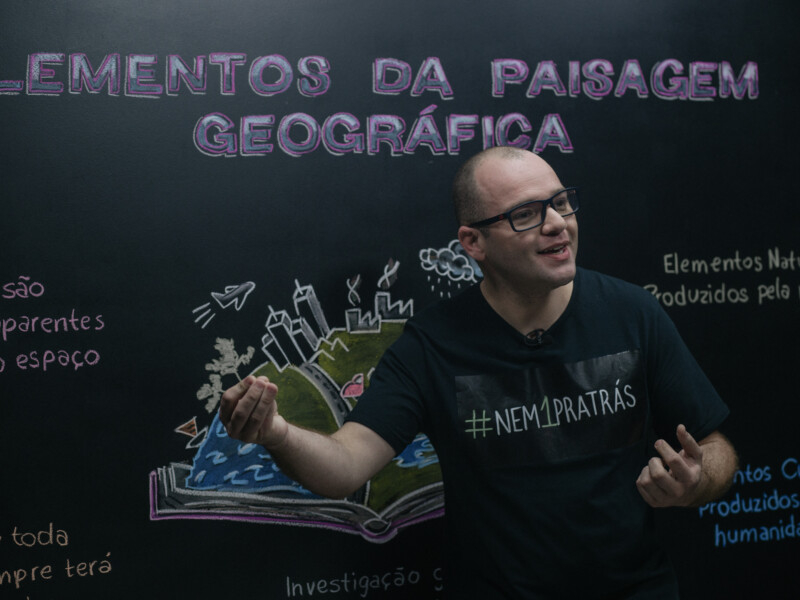Um professor em frente a um quadro negro gesticulando e falando algo. Ele é professor de geografia