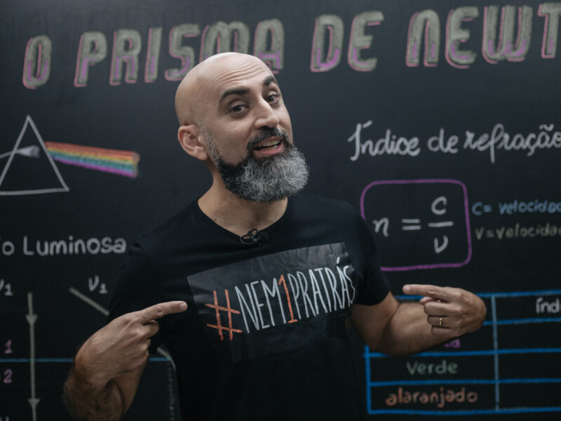 Professor careca, de barba volumosa e grisalha está posando para a foto em frente a um quadro negro com anotações sobre Física