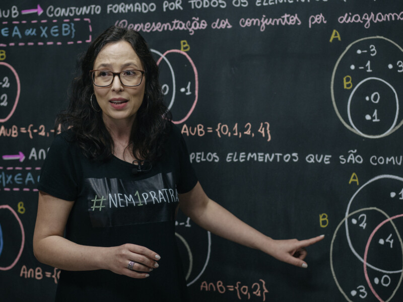 Professora branca de cabelos pretos, soltos e compridos está em frente a um quadro negro apontando para algo escrito no quadro e falando algo