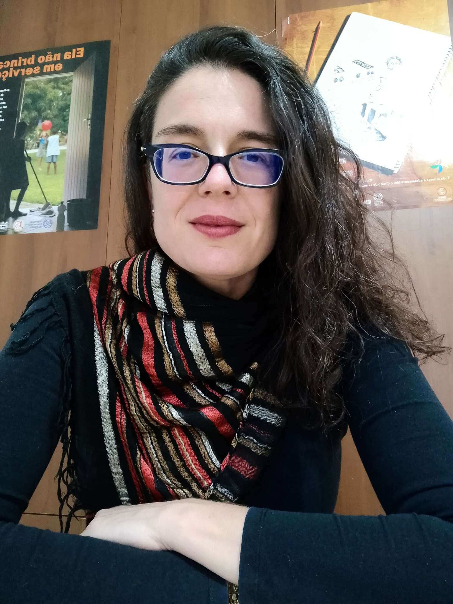 Uma mulher branca de óculos de grau, blusa preta de manga comprida, cachecol colorido e um cabelos soltos e escuros