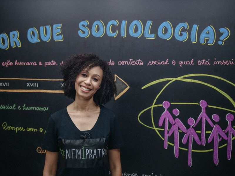 Professora negra de cabelos cacheados está em frente a um quadro negro com anotações sobre sociologia