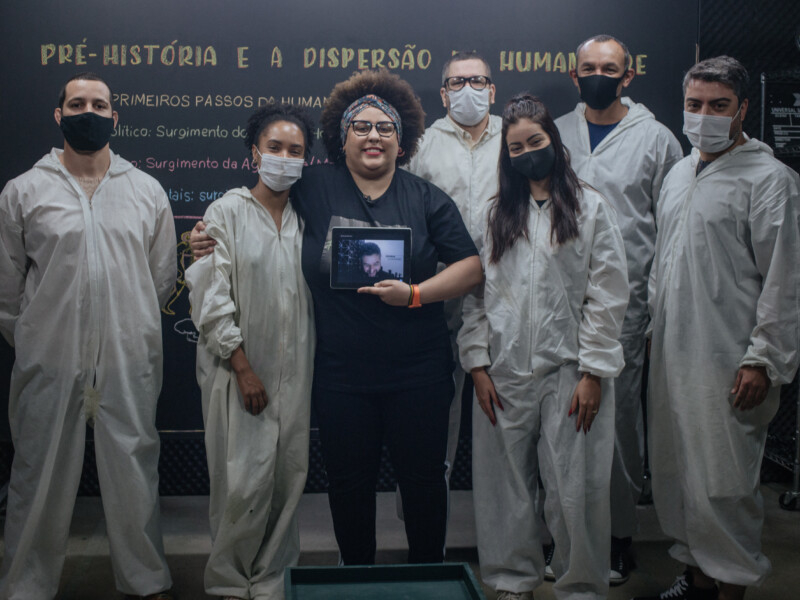 Seis pessoas estão posando para a foto comum quadro negro atrás delas. Cinco pessoas estão trajando uma mesma roupa branca e usando máscaras de prevenção à covid, já a mulher do centro da imagem está de preto e parece ser a professora