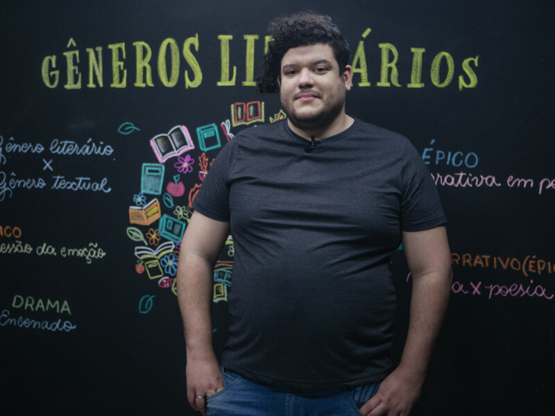 Professor negro, acima do peso e de cabelo cacheado está em frente a um quadro negro posando para a foto