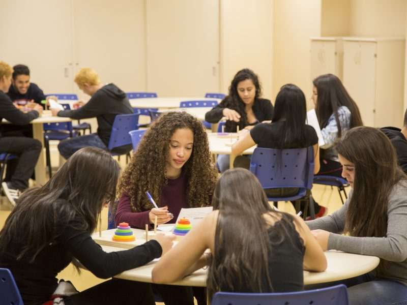 Quatro meninas sentadas em volta de uma mesa circular...ao fundo, outra mesa circular com três meninas sentadas ao redor